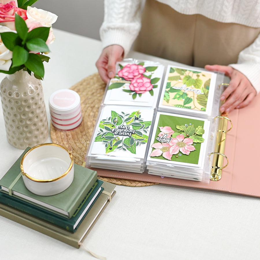 A person holds open a rouge binder filled with small square cards featuring colorful floral and botanical designs. The scene is styled on a white tabletop with a vase of flowers, stacked books, a small bowl, and a woven placemat. 