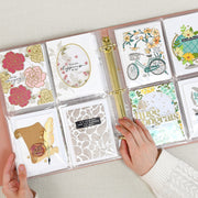 A top-down view of a clear plastic ring binder album with multiple illustrated craft cards displayed inside. Two hands are holding the binder open on a light fabric surface. 