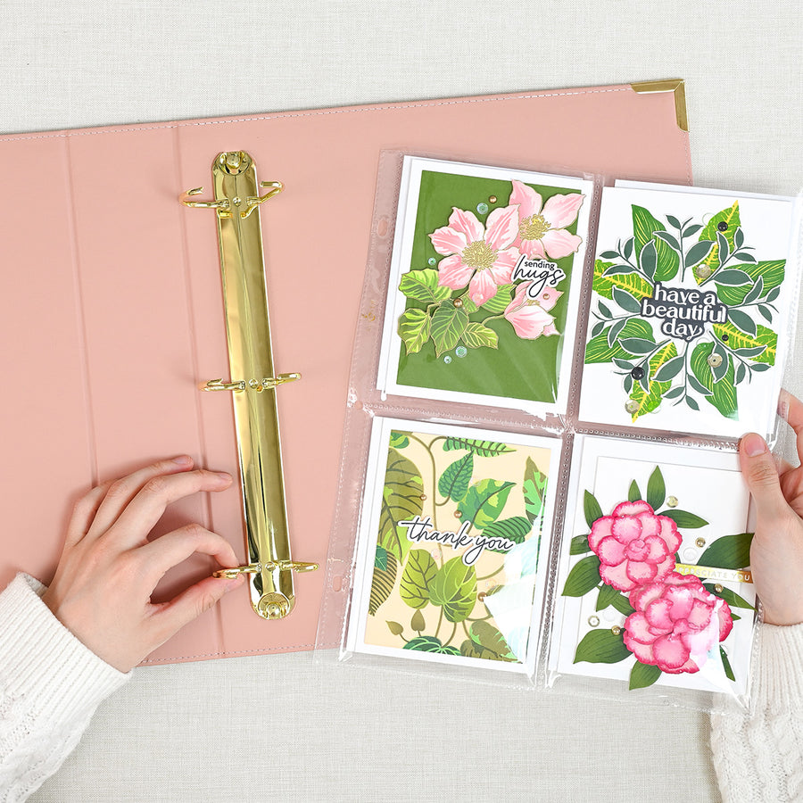 A rouge ring binder with a gold metal ring is shown open on a light fabric surface. Two hands are visible holding a clear plastic sleeve containing four decorative botanical cards with pink flowers and green leaves. 