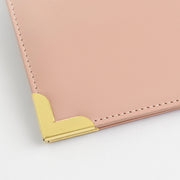 A close-up of a rouge leather folder or notebook cover with gold metal corner protectors and visible stitching, shown on a white background.