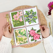 A top-down view of a person's hands holding an open rouge ring binder filled with four decorated card pages featuring floral and botanical designs, placed on a woven placemat with flowers and a pen nearby. 