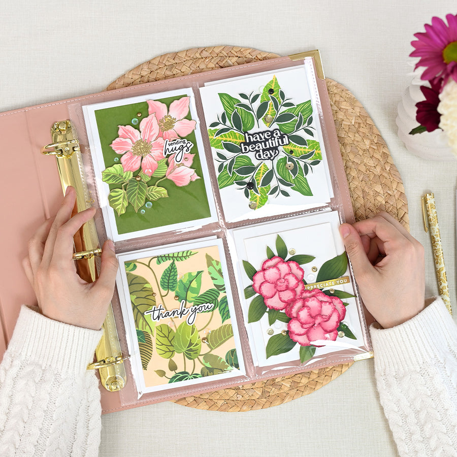 A top-down view of a person's hands holding an open rouge ring binder filled with four decorated card pages featuring floral and botanical designs, placed on a woven placemat with flowers and a pen nearby. 
