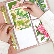 A top-down view of a person's hands organizing colorful botanical-themed cards in a rouge ring binder with clear pockets. The cards feature green leaves, pink flowers, and one card with 'thank you' written on it. 