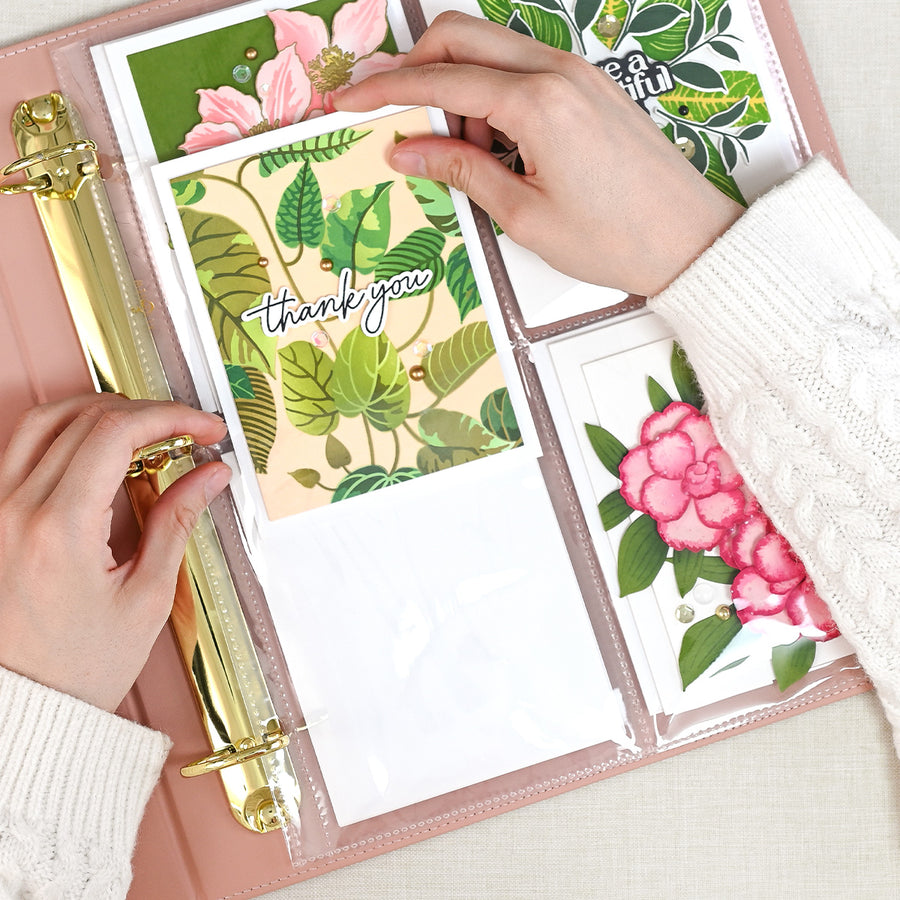A top-down view of a person's hands organizing colorful botanical-themed cards in a rouge ring binder with clear pockets. The cards feature green leaves, pink flowers, and one card with 'thank you' written on it. 