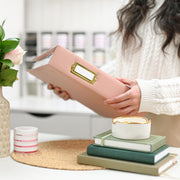 A person in a cream cable-knit sweater holds an ivory faux-leather binder with a gold-tone metal nameplate. The scene is set in a bright, modern room with a styled desk featuring stacked green and beige books, a small white candle with gold trim, and a pink rose in a textured vase. 