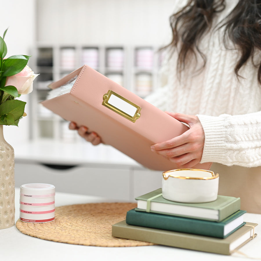 A person in a cream cable-knit sweater holds an ivory faux-leather binder with a gold-tone metal nameplate. The scene is set in a bright, modern room with a styled desk featuring stacked green and beige books, a small white candle with gold trim, and a pink rose in a textured vase. 