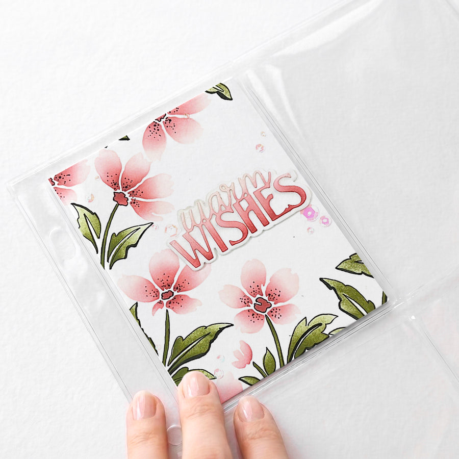 A hand holds a clear plastic sleeve containing a square card with pink floral artwork and the words 'again WISHES' in stylized lettering, photographed on a light neutral background.