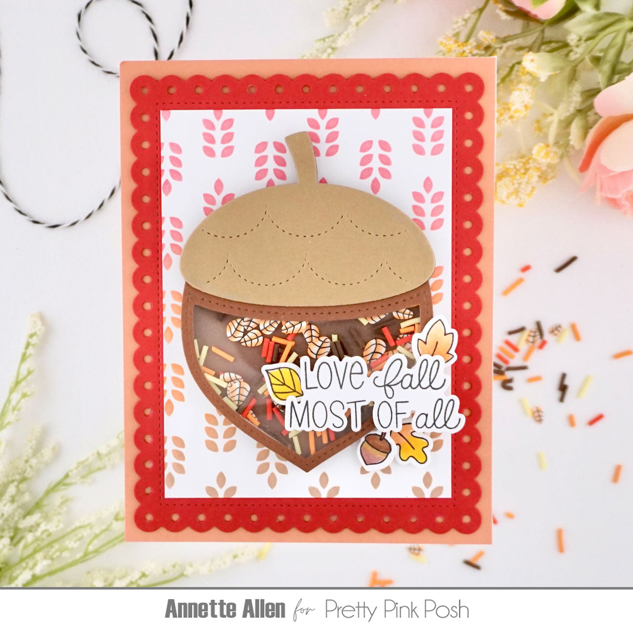 A handmade layered card featuring a die-cut acorn with a clear heart-shaped center filled with autumn candy and fall-themed embellishments. The card has a coral red scalloped border, a white background with pink leaf patterns, and the phrase 'Love Fall MOST OF all' in layered white and yellow die-cut elements.