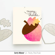 A handmade greeting card featuring a die-cut acorn design with pink glittery leaves and the words 'many thanks' in black script, displayed on a white background with metal die-cutting tools partially visible on both sides.