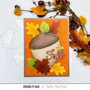 A handmade autumn-themed greeting card featuring an acorn and colorful fall leaves on an orange background, styled with decorative autumn branches on a white surface. The card is branded Debbie Frank for Pretty Pink Posh.