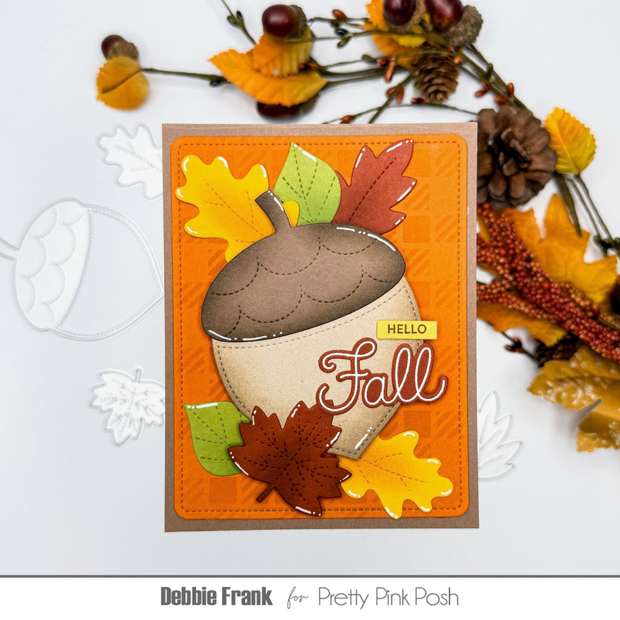 A handmade autumn-themed greeting card featuring an acorn and colorful fall leaves on an orange background, styled with decorative autumn branches on a white surface. The card is branded Debbie Frank for Pretty Pink Posh.