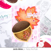 An autumn-themed card featuring a die-cut acorn with a gold foil shell, colorful confetti inside, and a layered orange and red maple leaf behind it with the text 'LOVE fall MOST OF all'. The card is displayed on a white background with other crafting supplies and stamps.