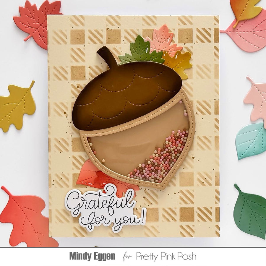A handmade autumn-themed card featuring a 3D acorn design with a textured brown cap, a beige body filled with multicolored sequins, and the phrase 'Grateful for you!' in white script. The card is displayed on a white background surrounded by colorful paper leaves, with the text 'Mindy Eggen for Pretty Pink Posh' at the bottom.