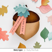 Acorn-shaped paper ornament with a brown top, tan body, and peach tags reading 'SUGAR FOR YOU' and 'TASTE UTTERLY', decorated with layered teal and peach leaves on a white background with scattered autumn leaves. Text at bottom reads 'Mindy Eggen for Pretty Pink Posh'.