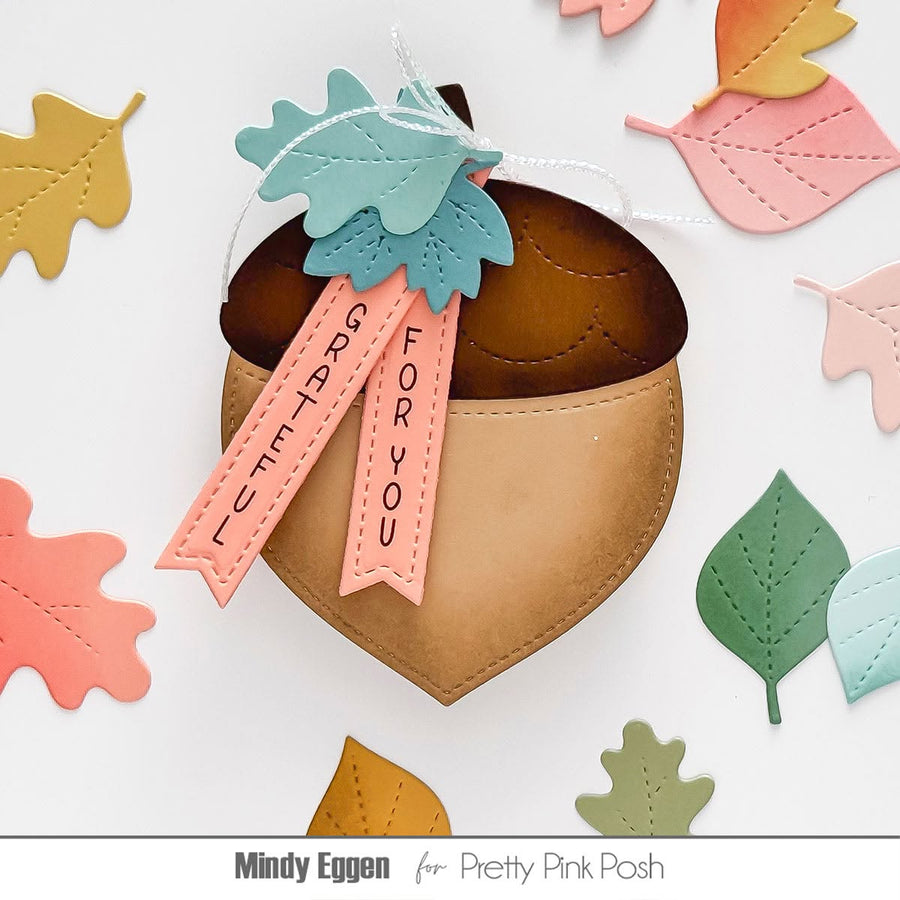 Acorn-shaped paper ornament with a brown top, tan body, and peach tags reading 'SUGAR FOR YOU' and 'TASTE UTTERLY', decorated with layered teal and peach leaves on a white background with scattered autumn leaves. Text at bottom reads 'Mindy Eggen for Pretty Pink Posh'.
