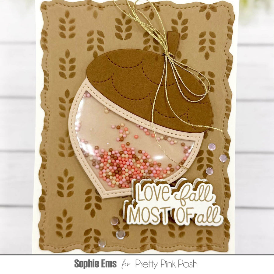 A handmade autumn-themed card featuring a die-cut acorn design with a clear window filled with pink and gold shimmery beads. The card has a scalloped tan border with stamped leaf patterns, a gold twine bow, and the text 'Love fall, MOST OF all' in white.