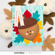 A handmade autumn-themed greeting card featuring a cute acorn face surrounded by colorful layered leaves on a light blue background. The card is styled on a white surface with decorative fall leaves, pumpkins, and pinecones.