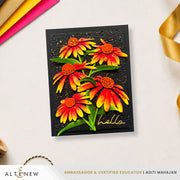 A handmade black greeting card featuring three dimensional yellow and red coneflowers with green leaves and gold glitter, with the word 'hello' written in gold foil script. The card is displayed on a white surface with colorful paper strips and a gold ribbon in the background. Altenew Craft-A-Flower: Coneflower Layering Die Set card ideas