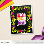 A handmade greeting card with a black textured background, decorated with a colorful floral border and the words 'HELLO SWEET friend' in layered pink, yellow, and purple lettering. The card is displayed on a light beige surface with craft paper and a purple ribbon in the corners.