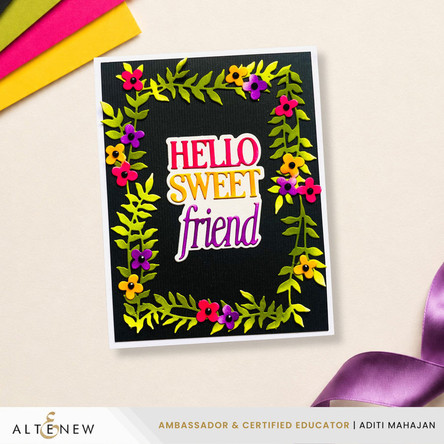 A handmade greeting card with a black textured background, decorated with a colorful floral border and the words 'HELLO SWEET friend' in layered pink, yellow, and purple lettering. The card is displayed on a light beige surface with craft paper and a purple ribbon in the corners.