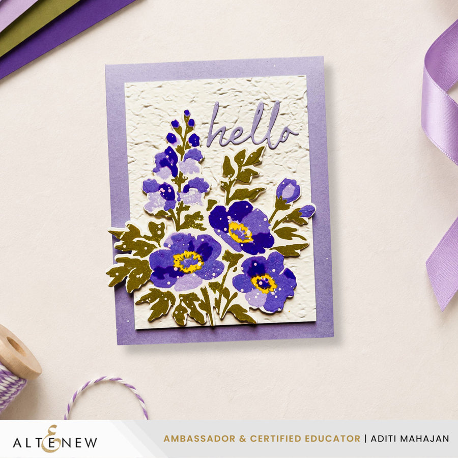 A handmade greeting card featuring purple and blue flowers with green leaves, a textured white center, and the word 'hello' in purple script, layered over a lavender border. The card is photographed on a light beige surface with ribbon and paper props around it. Altenew Craft Your Life Project Kit: Painterly Perfection card ideas