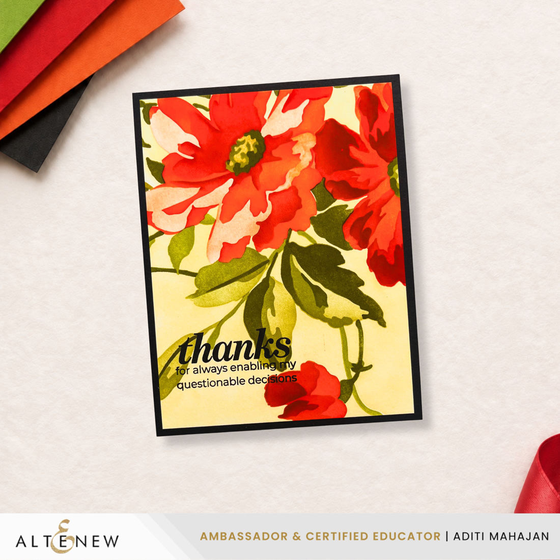 A handmade greeting card with a black border, featuring a watercolor-style floral design in red, orange, and green on a pale yellow background, with the text 'thanks for always enablingly questionable decisions.' It is displayed on a textured white surface with craft paper sheets in the corners.