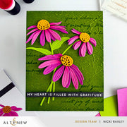 A handmade greeting card featuring three dimensional pink daisy flowers with yellow centers on a textured green background with cursive writing. The card has a black banner at the bottom with the text 'MY HEART IS FILLED WITH GRATITUDE'. Altenew Craft-A-Flower: Coneflower Layering Die Set card ideas