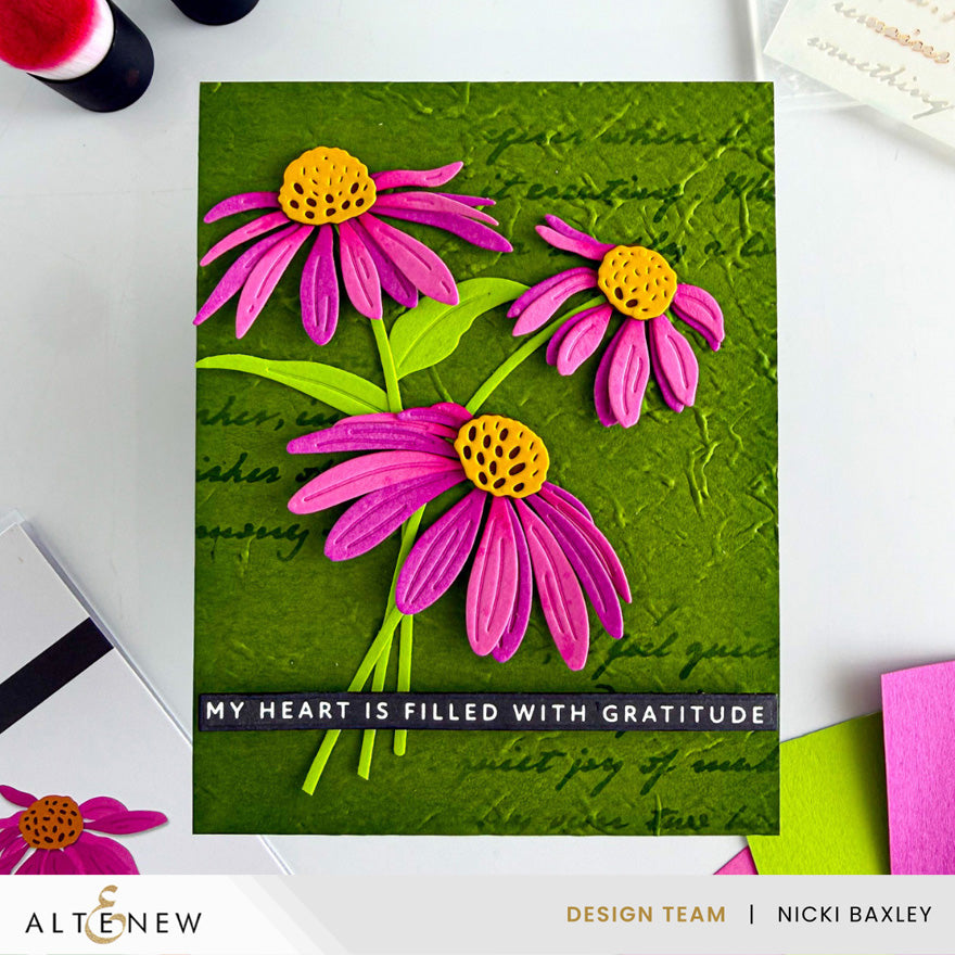 A handmade greeting card featuring three dimensional pink daisy flowers with yellow centers on a textured green background with cursive writing. The card has a black banner at the bottom with the text 'MY HEART IS FILLED WITH GRATITUDE'. Altenew Craft-A-Flower: Coneflower Layering Die Set card ideas