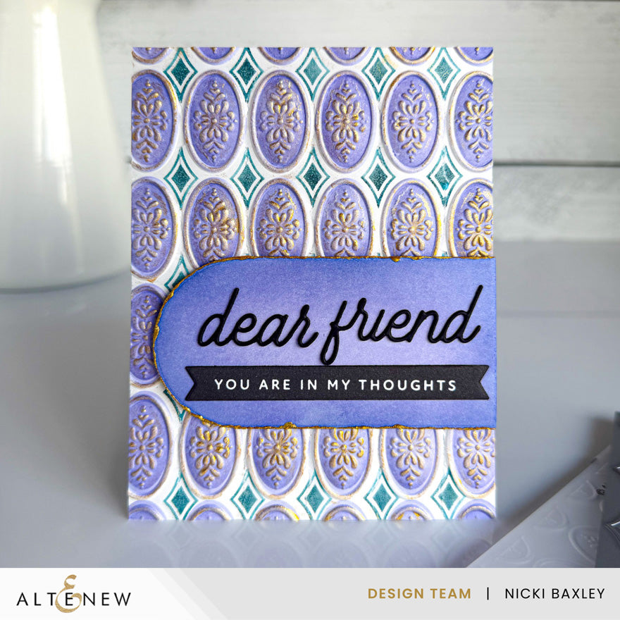 A handmade greeting card with a lavender and teal patterned background and a central purple banner reading 'dear friend YOU ARE IN MY THOUGHTS', photographed on a light surface with a blurred white vase in the background.