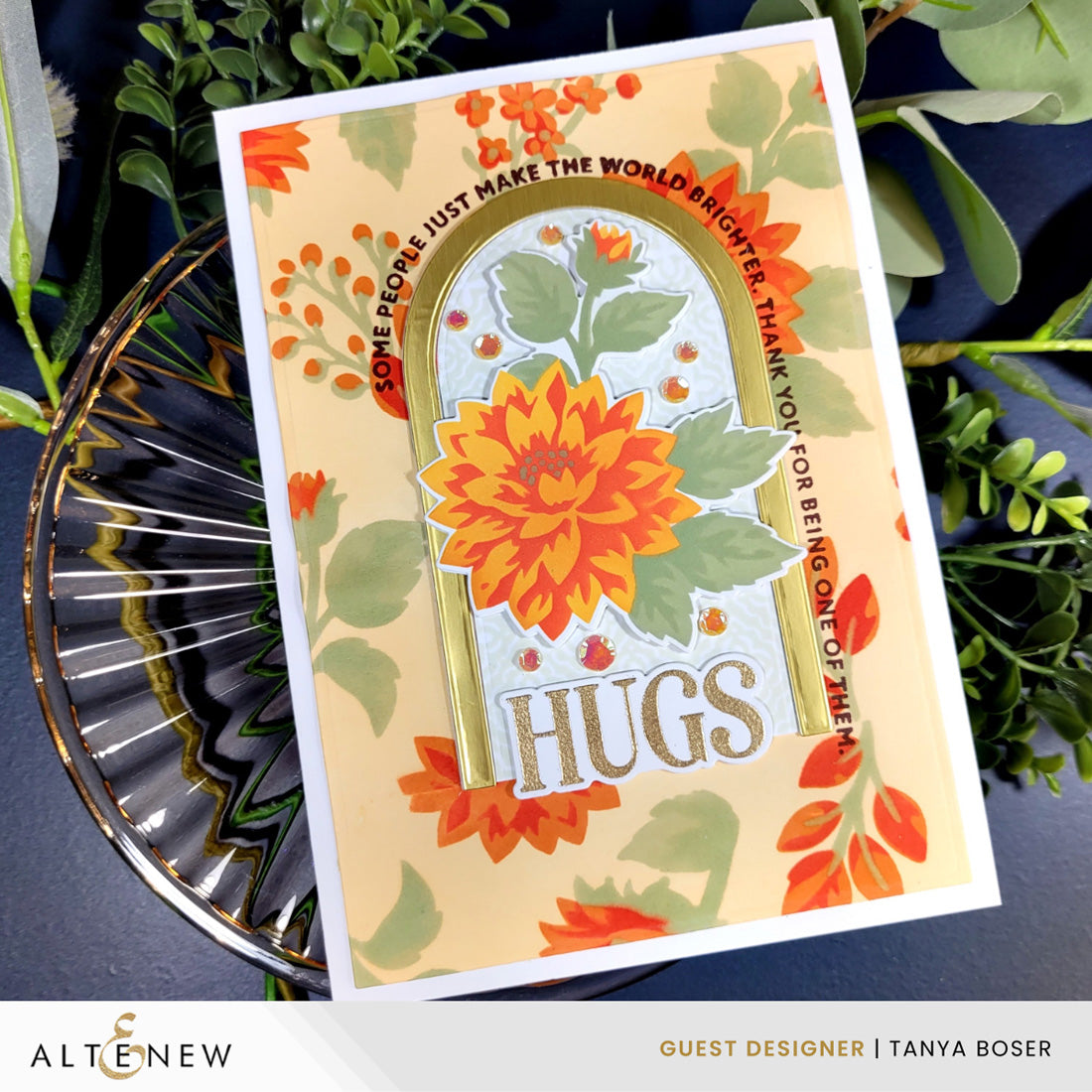 A handmade greeting card with a large orange dahlia flower and the word 'HUGS' in a gold arch, styled on a dark surface with green leaves and a glass dish. The card is branded with 'ALTERNOW' and 'GUEST DESIGNER | TANYA BOSER'.