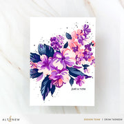 A vertical art print or card featuring a watercolor floral bouquet in purple, pink, and navy on a white background, with the text 'just a note' near the lower right. The print is photographed on a light marble surface, with ALTENEW branding and designer credits along the bottom. Altenew Build-A-Garden: Softly Sketched Blooms