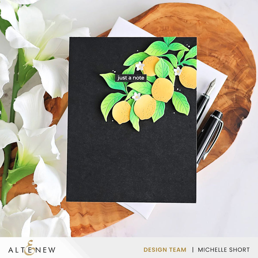 A black handmade greeting card featuring a 3D layered paper lemon design with green leaves and the text 'just a note', styled on a wooden slice with white flowers and pens. Altenew Creative Cuts: Lemon Tree Layering Die Set card ideas