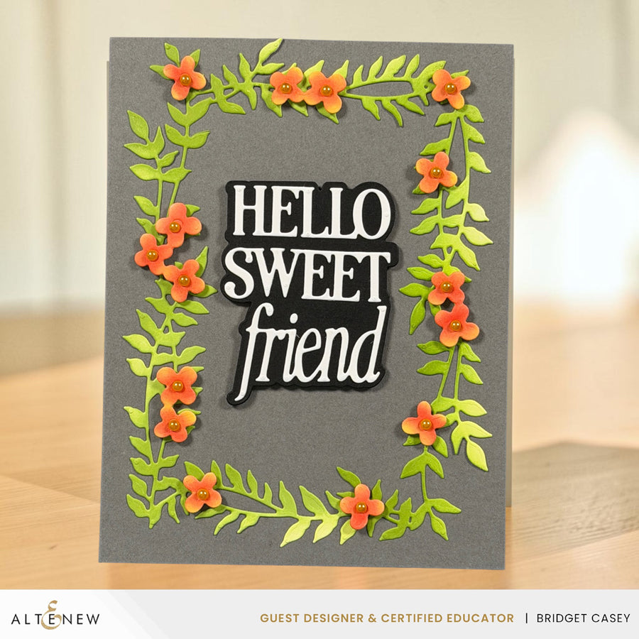 A handmade greeting card with a dark gray background, featuring a raised black and white 'HELLO SWEET friend' text element surrounded by layered green leaves and orange flowers, displayed on a wooden surface.