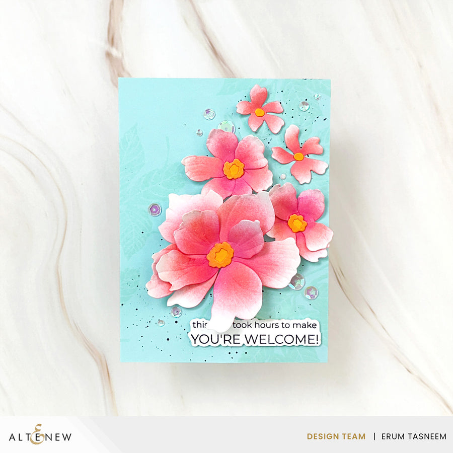 A handmade greeting card featuring layered pink and peach flowers with yellow centers on a light blue background, with the text 'YOU'RE WELCOME!' and a small decorative quote. The card is photographed on a white marble surface, with the Altenew logo and designer credit at the bottom.