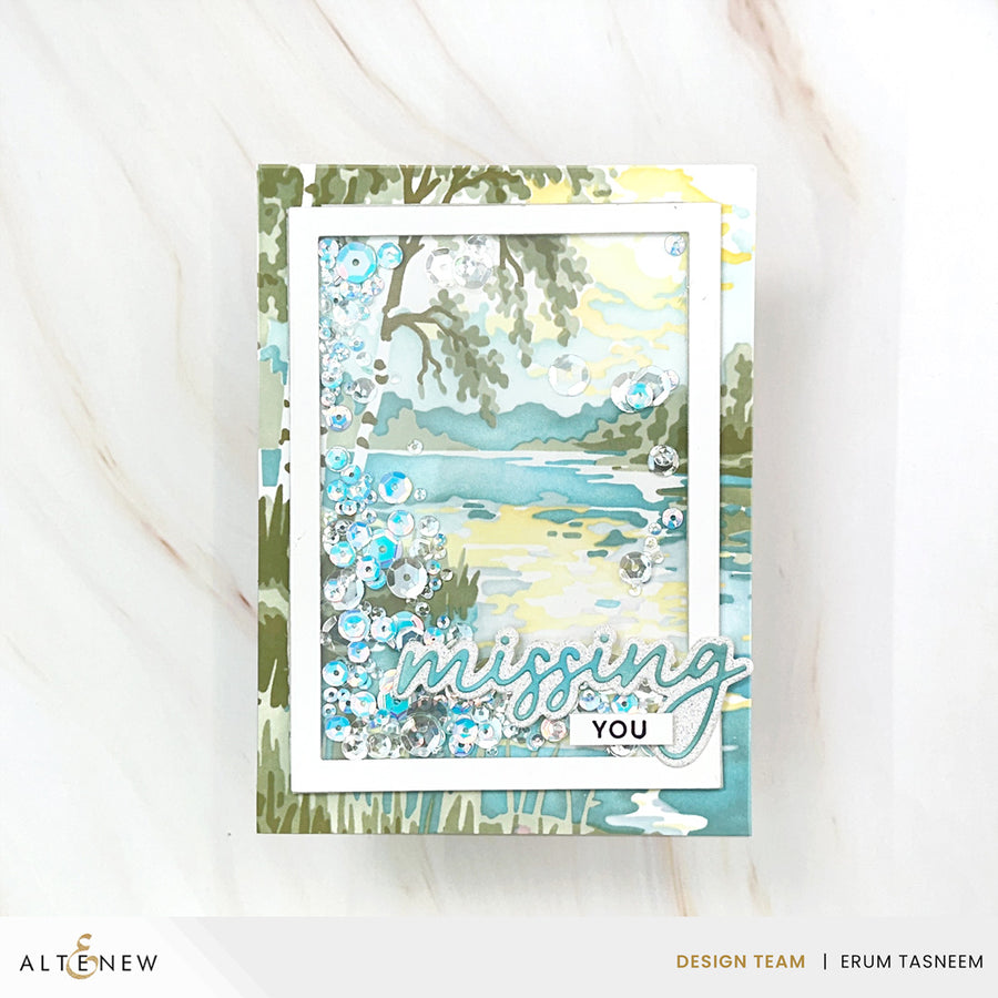 A handmade layered card featuring a watercolor-style lake landscape with blue and green tones, floral embellishments, and the words 'missing YOU' in script and block lettering, displayed on a light marble surface. Altenew Craft Your Life Project Kit: Painterly Perfection card ideas