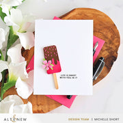 A white handmade greeting card featuring a popsicle illustration with pink frosting, chocolate brown topping, and a small pink flower, placed on a wooden slice with pink paper, pens, and white flowers. The card has the text 'LIFE IS SWEET WITH YOU IN IT'. Altenew Mini Delight: Sweet Popsicles Stamp & Die Set card ideas