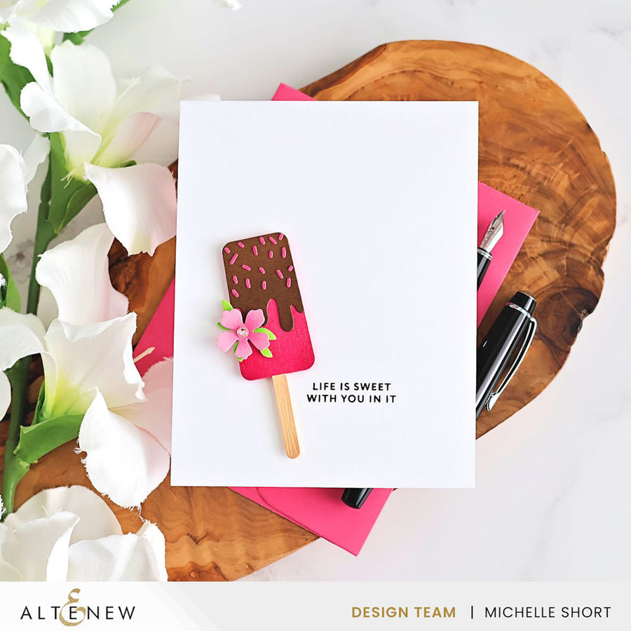 A white handmade greeting card featuring a popsicle illustration with pink frosting, chocolate brown topping, and a small pink flower, placed on a wooden slice with pink paper, pens, and white flowers. The card has the text 'LIFE IS SWEET WITH YOU IN IT'. Altenew Mini Delight: Sweet Popsicles Stamp & Die Set card ideas