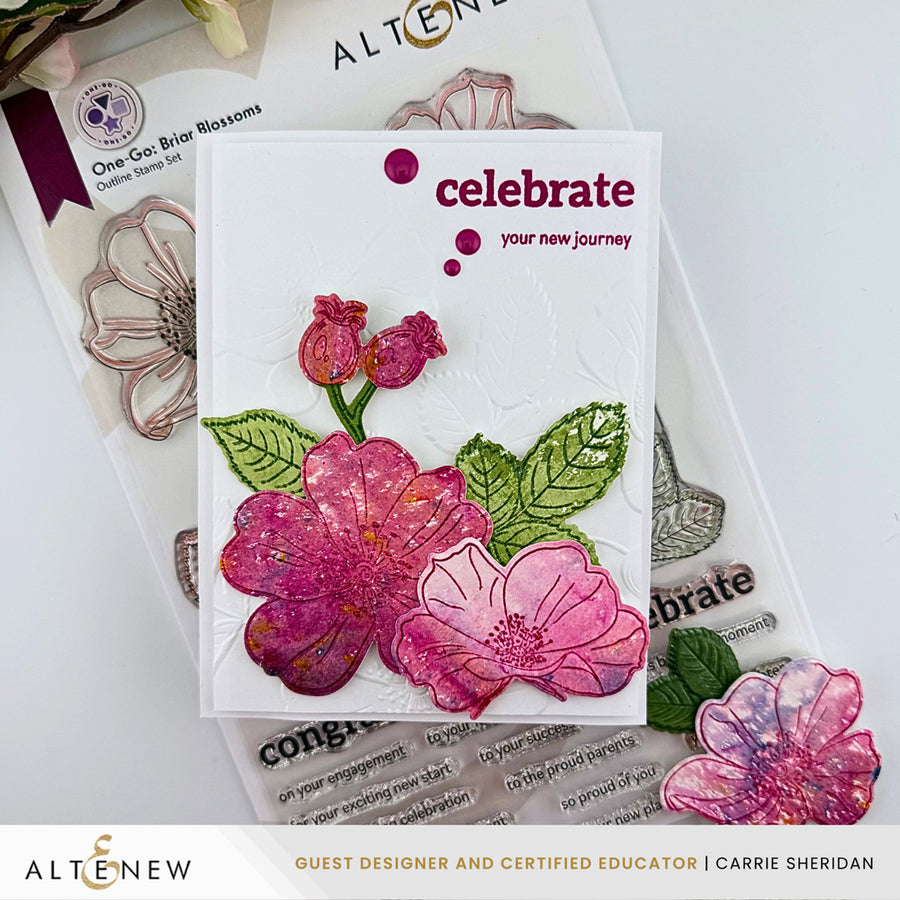 A handmade greeting card featuring pink and magenta embossed flowers with green leaves on a white background, with the words 'celebrate your new journey' printed in dark pink. The card is displayed on top of Altenew crafting materials and stamps. Altenew One-Go: Briar Blossoms card ideas