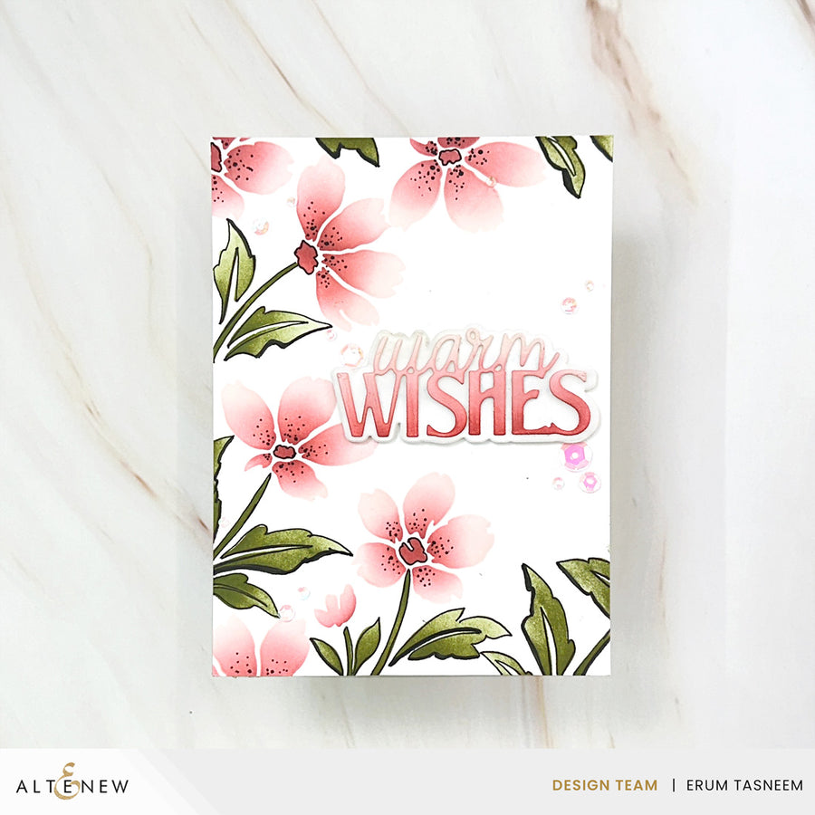 A handmade greeting card with a white background, featuring pink watercolor-style flowers with green leaves and the words 'warm WISHES' in pink script and block lettering. The card is photographed flat on a light marble surface, with brand text 'ALT&ENew' and 'DESIGN TEAM | ERUM TASNEEM' visible at the bottom. Altenew Painted Petals Stencil Set (3 in 1) card ideas