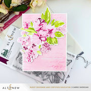 A handmade greeting card with layered pink and green floral die-cut stickers on a light pink background, styled on top of clear stamp blocks and metal stamp dies. The card has the text 'wishing you a lovely day' and the brand name 'ALTINEEW' is visible on the packaging and in the lower banner. Altenew Build-A-Garden: Softly Sketched Blooms