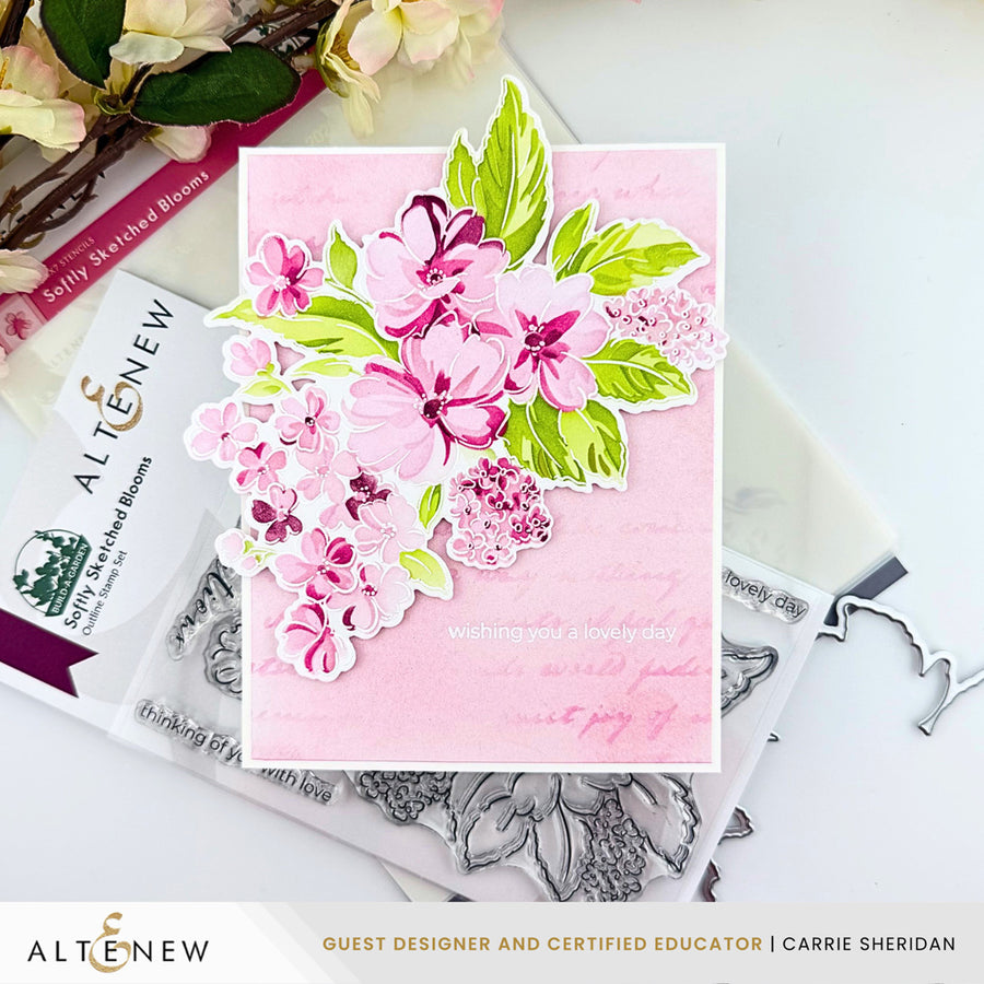 A handmade greeting card with layered pink and green floral die-cut stickers on a light pink background, styled on top of clear stamp blocks and metal stamp dies. The card has the text 'wishing you a lovely day' and the brand name 'ALTINEEW' is visible on the packaging and in the lower banner. Altenew Build-A-Garden: Softly Sketched Blooms