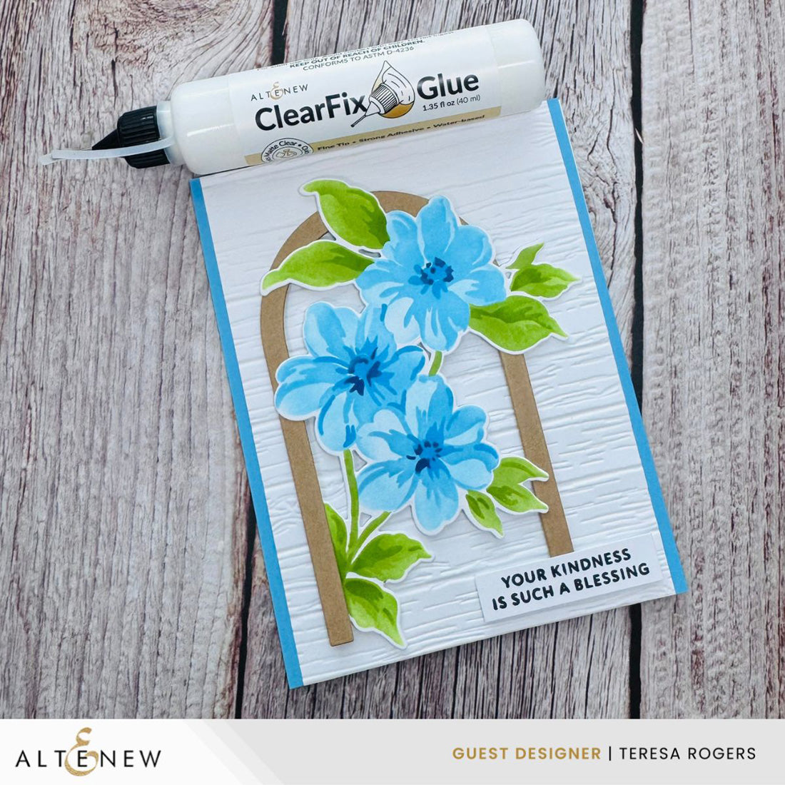 An Altenew ClearFix Glue bottle is placed diagonally over a handmade greeting card on a rustic wood background. The card features layered blue flowers with green leaves on a textured white background with a tan arch, and the text 'YOUR KINDNESS IS SUCH A BLESSING'.