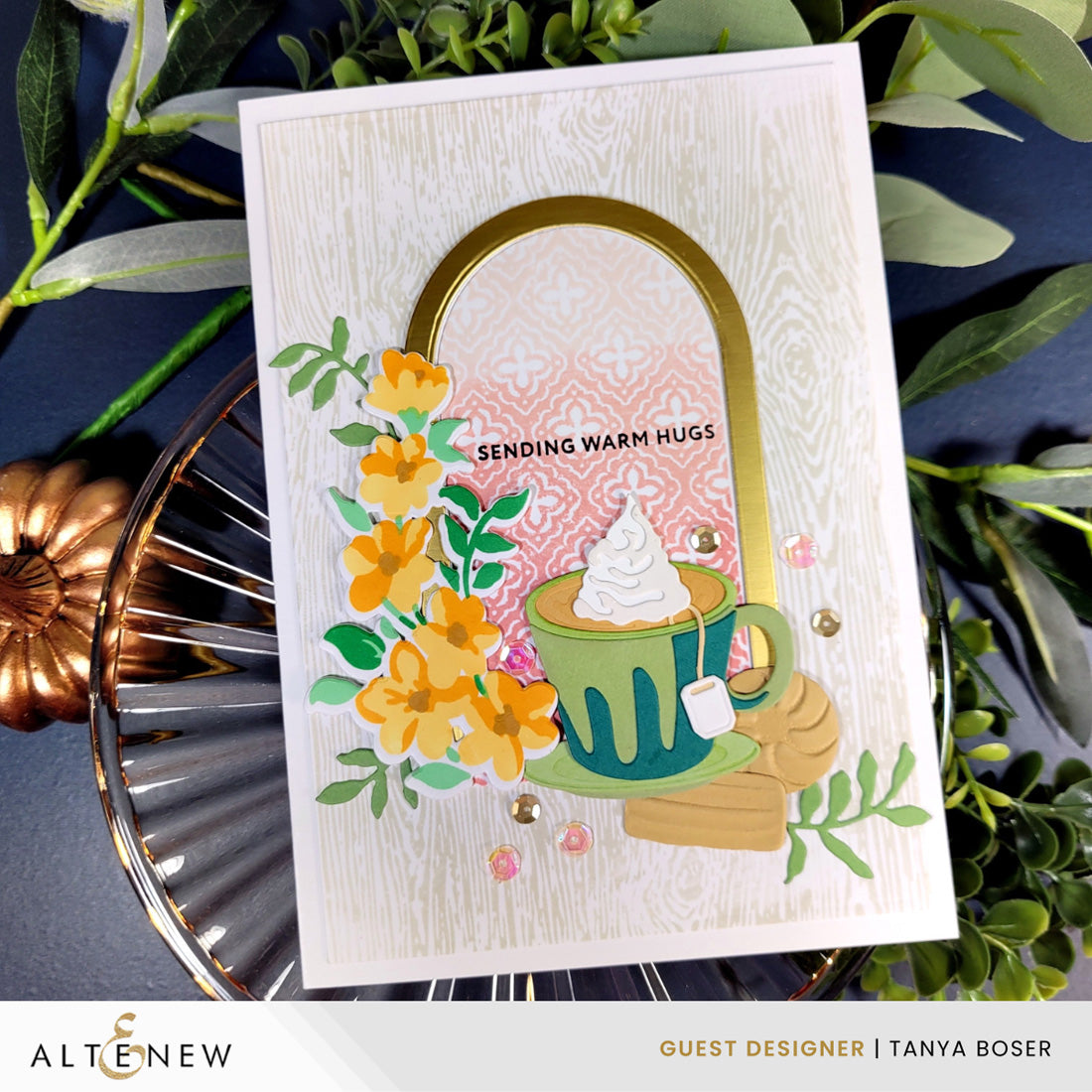 A handmade greeting card featuring a green mug with whipped cream, yellow flowers, and the text 'SENDING WARM HUGS', styled on a dark blue surface with green leaves and a copper pumpkin. The card is branded with 'ALTENEW' and 'GUEST DESIGNER | TANYA BOSER' at the bottom.