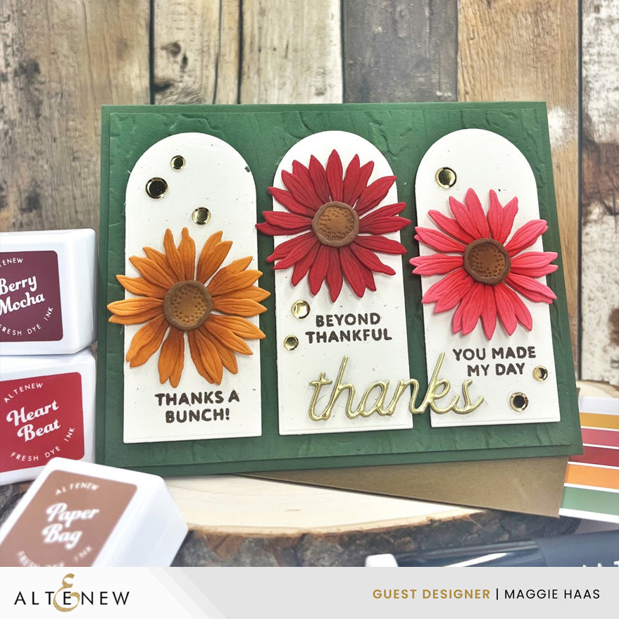 A handmade greeting card displayed on a wooden surface with Alteneew ink pads nearby. The card has a dark green background with three white arch-shaped panels featuring colorful paper flowers in orange, red, and pink, plus gold foil details and printed thank-you messages.