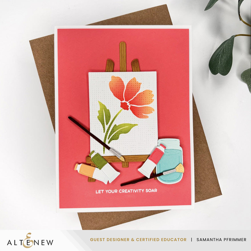 A handmade greeting card featuring a colorful illustration of a flower on an easel with paintbrushes, paint tubes, and a paint jar. The card has a coral pink background and is displayed on top of a brown kraft envelope on a white surface with a green leaf in the corner. Altenew Painted Petals Stencil Set (3 in 1) card ideas