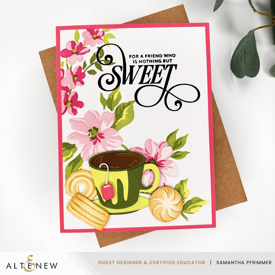 A greeting card with a pink border and illustrated flowers, a green teacup, cookies, and the word 'SWEET' in black script, displayed on top of a brown envelope against a white background with a small green leaf in the corner.