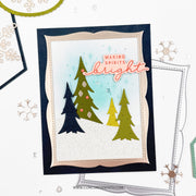 A handmade holiday greeting card featuring three stylized Christmas trees in navy, olive green, and yellow-green on a white glitter background with a light blue wash. The card has scalloped beige and navy borders, the phrase 'MAKING SPIRITS bright' in pink and orange, and is photographed on a white surface with craft tools and die-cut paper around it.