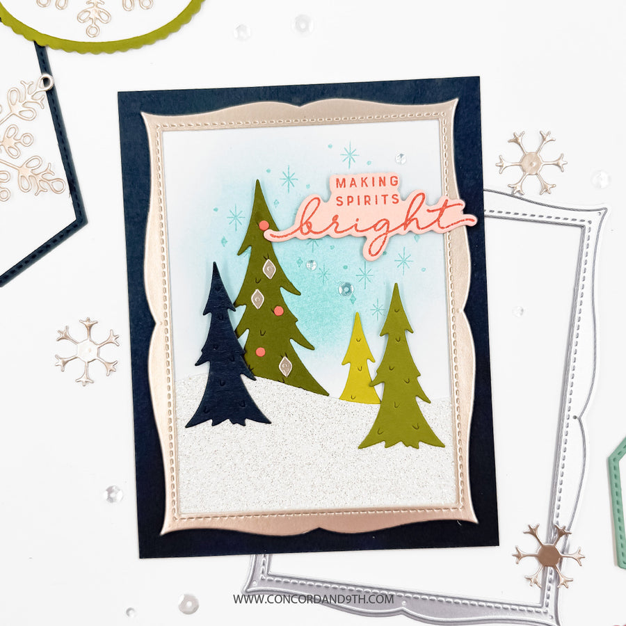 A handmade holiday greeting card featuring three stylized Christmas trees in navy, olive green, and yellow-green on a white glitter background with a light blue wash. The card has scalloped beige and navy borders, the phrase 'MAKING SPIRITS bright' in pink and orange, and is photographed on a white surface with craft tools and die-cut paper around it.