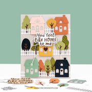A handmade layered card featuring a whimsical autumn village scene with houses, trees, and a central die-cut phrase reading 'You feel like HOME to me.' The card is displayed upright on a white surface against a pale green background, with small cutout pieces and a website watermark visible around it.