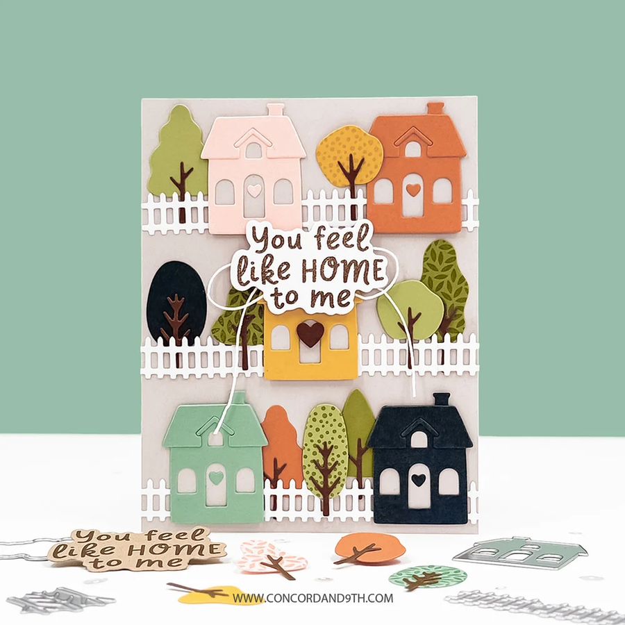 A handmade layered card featuring a whimsical autumn village scene with houses, trees, and a central die-cut phrase reading 'You feel like HOME to me.' The card is displayed upright on a white surface against a pale green background, with small cutout pieces and a website watermark visible around it.